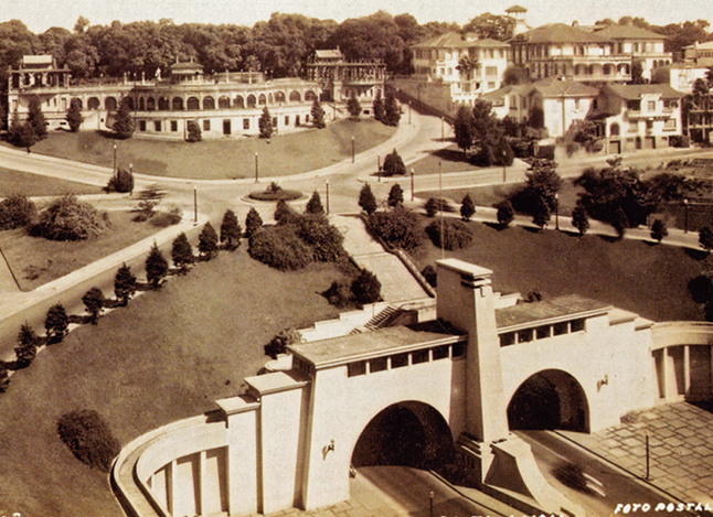 Fotografia em tons de sépia. Vista geral de local com dois arcos à frente, ao fundo, local com grama e árvores. Em segundo plano, rua de formato arredondado, construções, casas à direita e mais atrás, árvores.