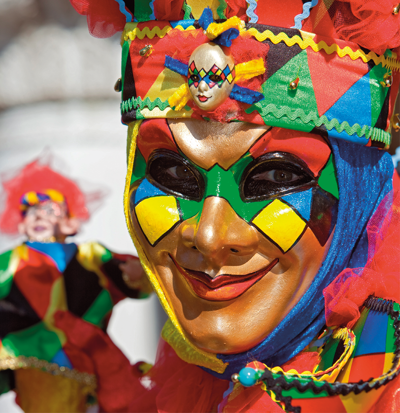 Fotografia. Rosto de uma pessoa de olhos pretos, voltado para a direita. Ela usa uma máscara dourada decorada com losangos coloridos em vermelho, verde, azul e amarelo. Embaixo do queixo, um pano em amarelo e azul contorna o rosto. A pessoa usa um adereço de cabeça nas mesmas cores. A máscara tem um sorriso com lábios em vermelho. Ao fundo, à esquerda, um boneco desfocado usando roupa também de losangos coloridos