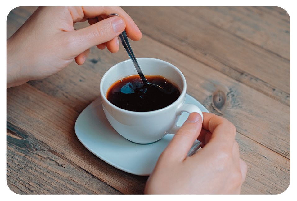 Fotografia. Duas mãos de uma pessoa, com a mão esquerda segurando uma colher pequena cinza sobre xícara branca com café dentro. A mão direita segurando alça da xícara. Debaixo dela, pires branco, sobre mesa de madeira em marrom.