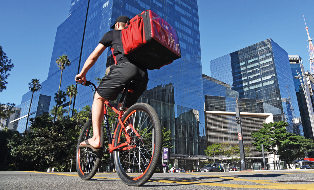 Fotografia. Vista de baixo para cima. À esquerda, rua cinza com um homem sobre uma bicicleta vermelha. Ele usa boné, camiseta, bermuda e par de chinelos em preto, com uma bolsa grande térmica nas costas em vermelho. Ao fundo, local com vários prédios espelhados em azul. Perto deles, árvores de folhas verdes. No alto, céu em azul-claro e sem nuvens.