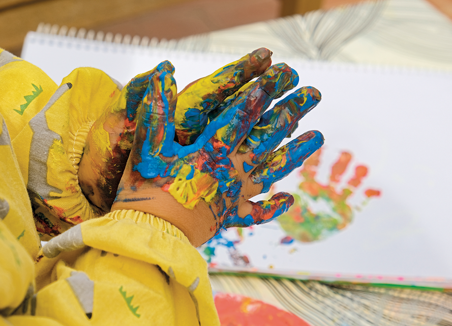 Fotografia. Vista geral de uma criança, com blusa de mangas compridas em amarelo, com as mãos unidas, cobertas com tintas em azul, vermelho e tons de amarelo. Ao fundo, papeis brancos com marca de uma mão em tons de vermelho, amarelo e verde.