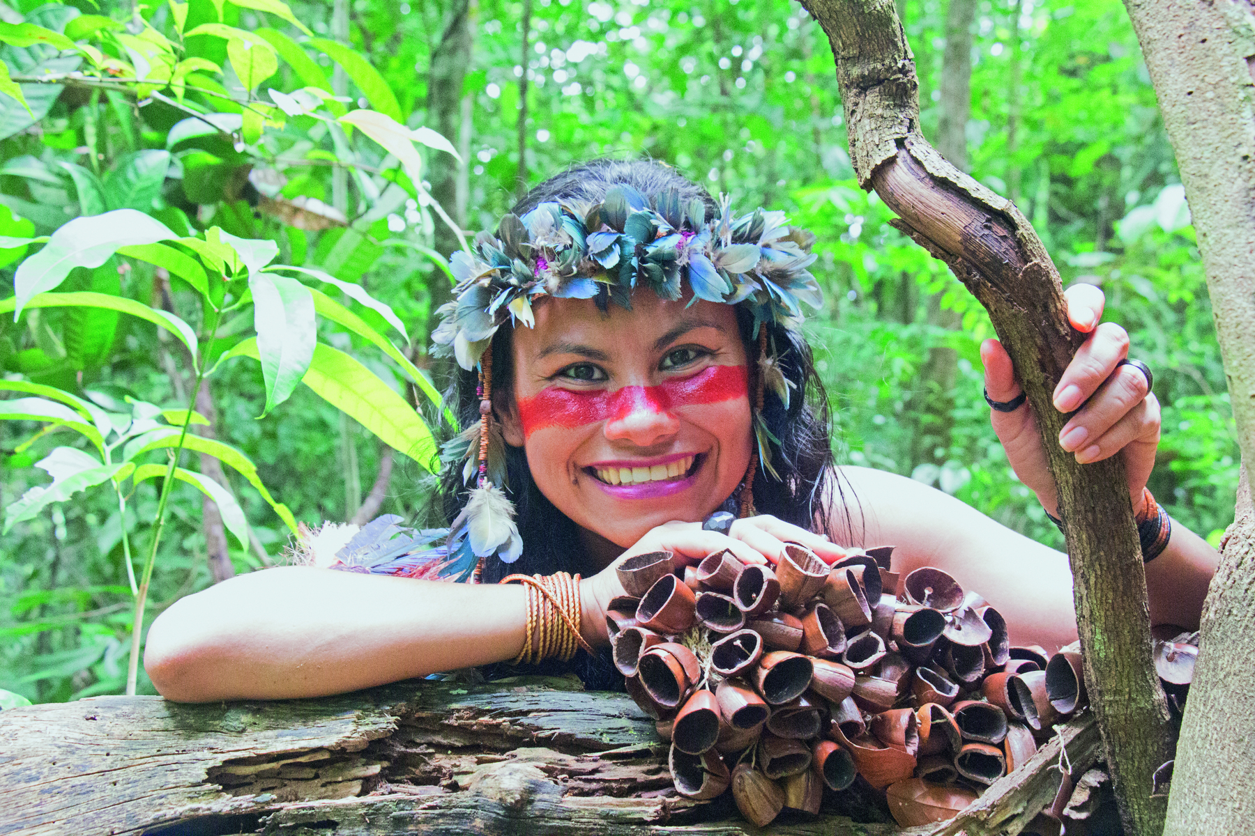 Fotografia. Uma mulher vista dos ombros para cima, de frente para tronco de árvore em marrom. Ela tem pinturas em vermelho nas bochechas, cabelos longos escuros e sobre a cabeça, uma coroa de folhas em verde. Ela olha para frente sorrindo, com o braço direito e cotovelo no tronco a frente e segurando um tronco fino na mão esquerda, na vertical, em marrom. Em segundo plano, arvores e vegetação densa.