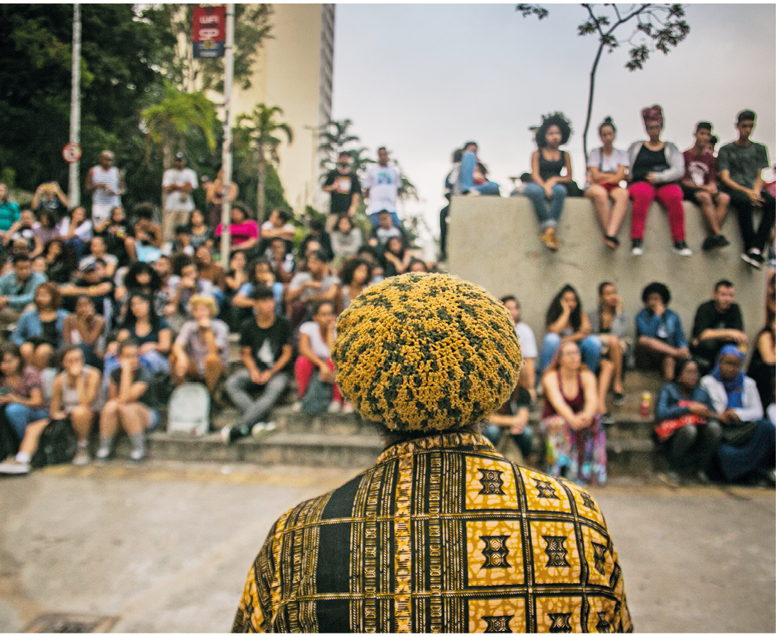 Fotografia. À frente, um homem visto de costas, da cintura para cima. Ele tem uma boina arredondada em tons de amarelo com partes em verde-escuro. Usa blusa de mangas compridas em tons de amarelo e em tons de verde-escuro. Ao fundo, dezenas de jovens juntos, à esquerda, sentados sobre o chão e à direita, sobre um muro em cinza. Em segundo plano, árvores de folhas em verde e céu acima em tons de cinza.