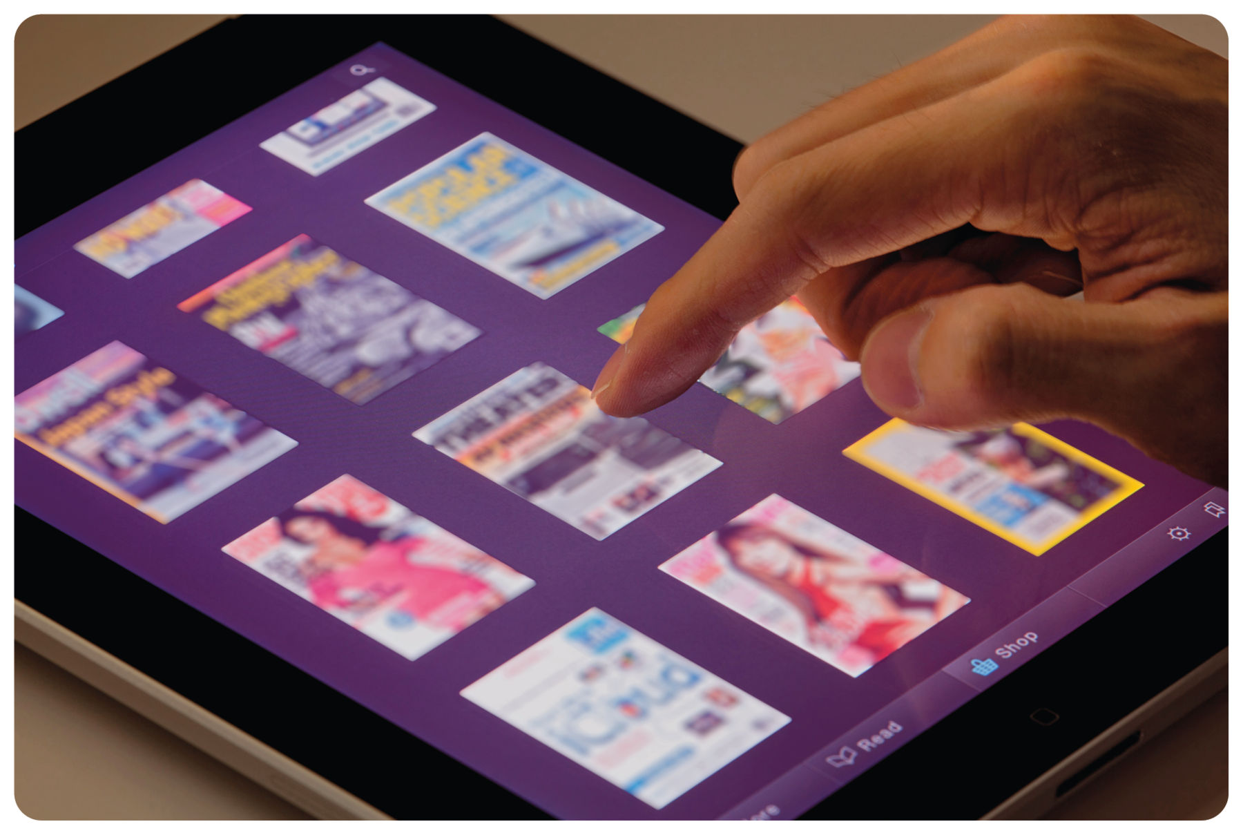 Fotografia. Um tablet retangular preto, sobre uma base marrom. Sobre o tablet, mão de uma pessoa com o dedo indicador clicando sobre foto de uma capa de revista. Ao redor, outras duas colunas na vertical com capas diversas.