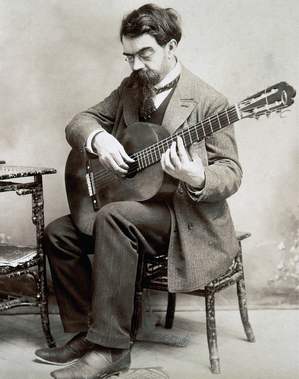 Fotografia em preto e branco. Um homem sentado sobre uma cadeira, segurando um violão nas mãos. Ele tem cabelos, barba e bigode escuros, com camisa branca por dentro, terno e sapatos escuros, olhando para baixo e mãos sobre violão. Mais à esquerda, vista parcial de mesa.