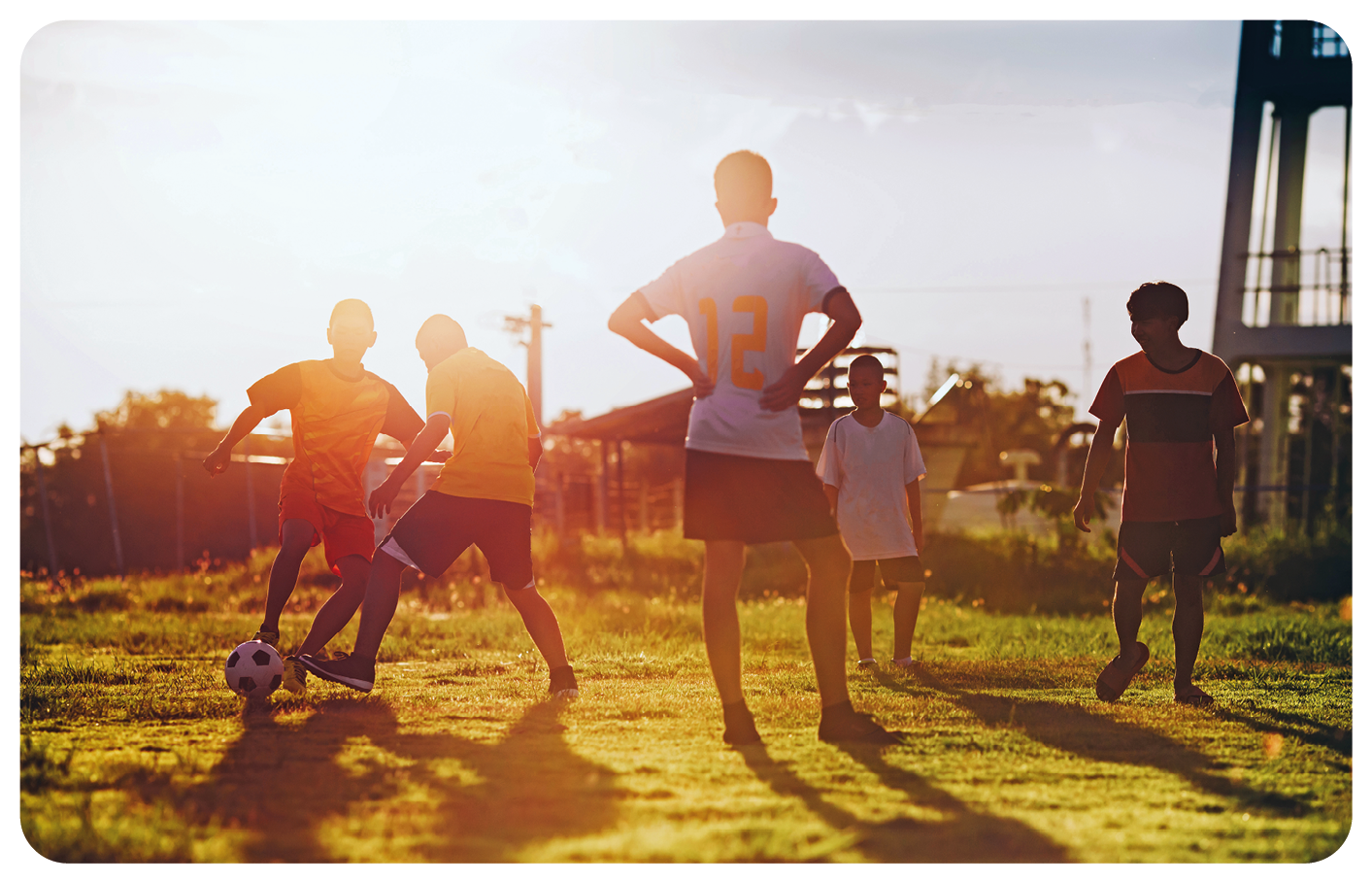 Fotografia. Cinco meninos em pé, jogando futebol com bola branca com detalhes em preto, em local com vegetação rasteira. Em segundo plano, vegetação, árvores de folhas verdes. Mais à direita, vista parcial de uma torre na vertical. No alto, céu em cinza e luz solar à esquerda.