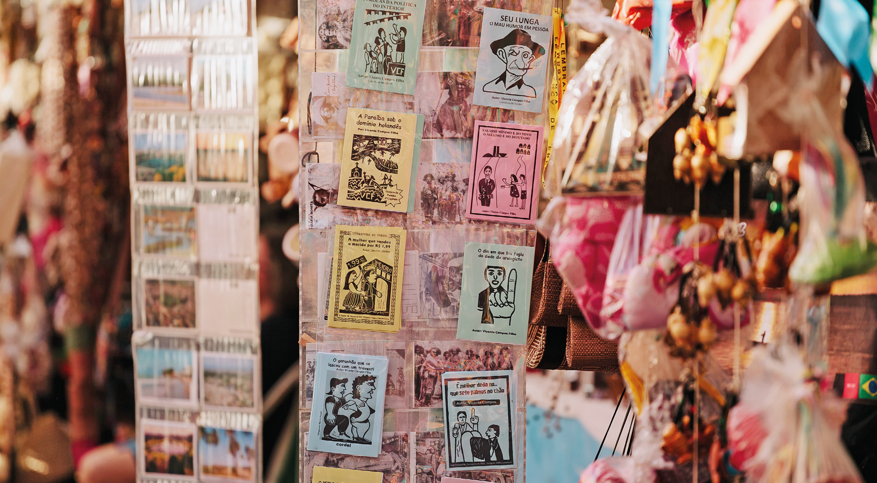 Fotografia. Vista geral de local com cordéis pendurados na vertical. Os livros têm folhas de cores diferentes, em azul, amarelo e rosa, com ilustrações em preto com xilogravura. Eles estão organizados na vertical.