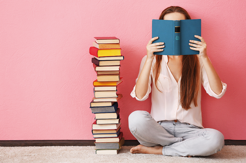 Fotografia. De frente para parede de cor rosa, uma menina sentada. Ela tem cabelos longos castanhos, de blusa de cor branca até os cotovelos, com calça cinza e descalça. A menina segura nas mãos, um livro de capa em azul, aberto, próximo do rosto. À esquerda, vários livros um sobre o outro, de capas coloridas, empilhados.