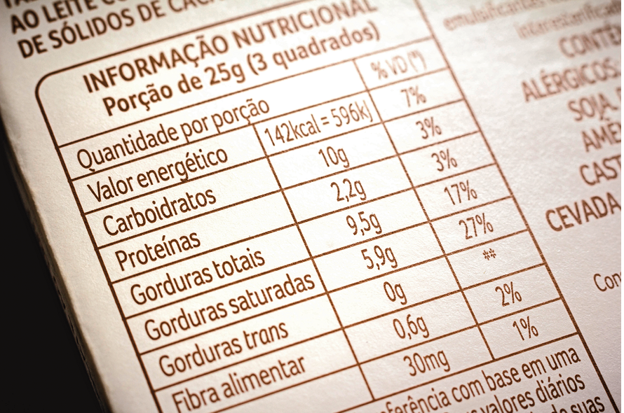 Reprodução de trecho de rótulo. Vista parcial de rótulo de cor branca e tabela de cor preta. Texto: Informação Nutricional Porção de 25g (3 quadrados) Quantidade por porção % VD(*) Valor energético 142 Kcal = 596 K – 7% VD(*) Carboidratos 10g 3% VD(*) Proteínas 2,2g 3% VD(*) Gorduras totais 9,5g 17% VD(*) Gorduras saturadas 5,9g 27% VD(*) Gorduras trans 0g ** VD(*) Fibra alimentar 0,6 2% VD(*) 30 mg 1% Na parte inferior, vista parcial de texto.