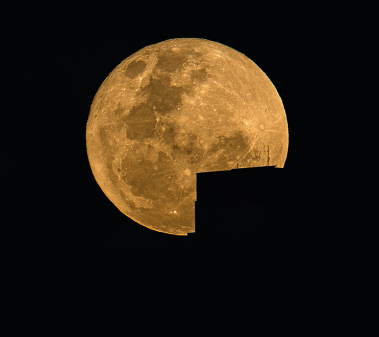 Fotografia. Vista de céu noturno em preto e, ao centro, lua redonda em bege. Na ponta inferior, à direita, sombra de ponta de telhado de formato triangular, em preto, sobre a lua.