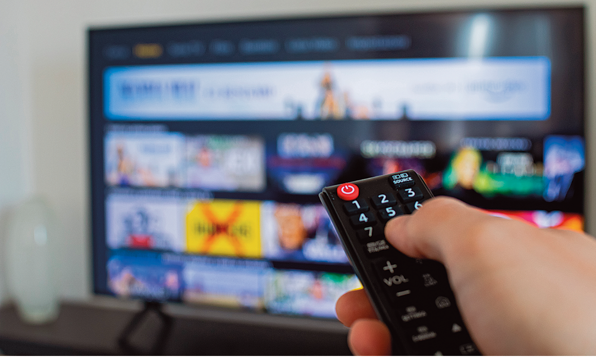 Fotografia. Mão de uma pessoa segurando um controle remoto preto com vários botões. Ao fundo, tela de TV na horizontal em preto com imagens embaçadas.