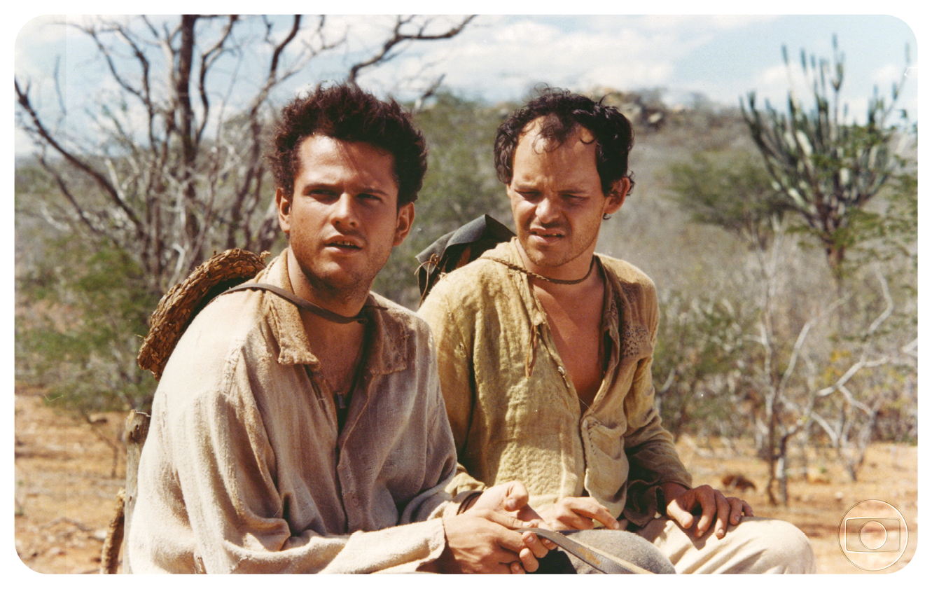 Cena de filme. Dois personagens sentados sobre local com solo em bege e vegetação seca, em tons de marrom e verde. À esquerda, homem de cabelos castanhos, de blusa de mangas compridas fina, em bege-claro. Ele olha para frente, tem chapéu bege, preso com um cordão preto ao redor do pescoço e mãos sobre os joelhos. À direita, homem um pouco calvo e cabelos escuros, vestido de camisa de cor bege-escuro e calça em bege-claro. Olha para frente e tem chapéu cinza preso com um cordão preto ao redor do pescoço. No alto, céu em azul-claro e nuvens brancas.