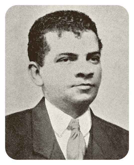 Fotografia em preto e branco. Um homem visto dos ombros para cima, de cabelos escuros, encaracolados, com camisa de gola clara, gravata clara e terno escuro.