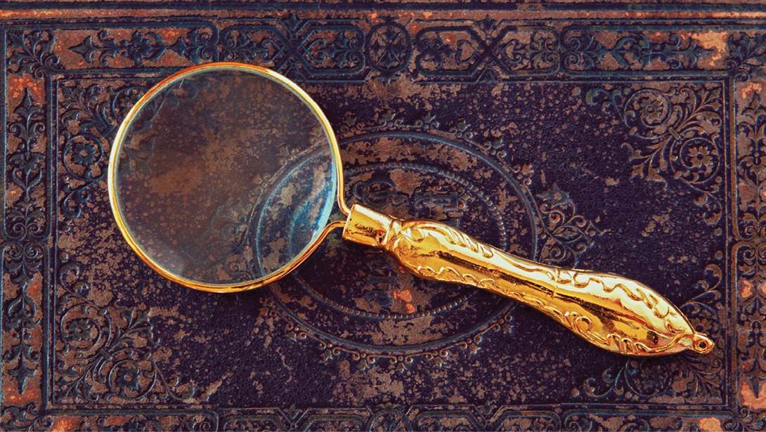 Fotografia. Sobre base na horizontal de madeira em marrom-escuro, com partes em marrom-claro, uma lupa grande e dourada, com a parte superior redonda e vidro transparente.