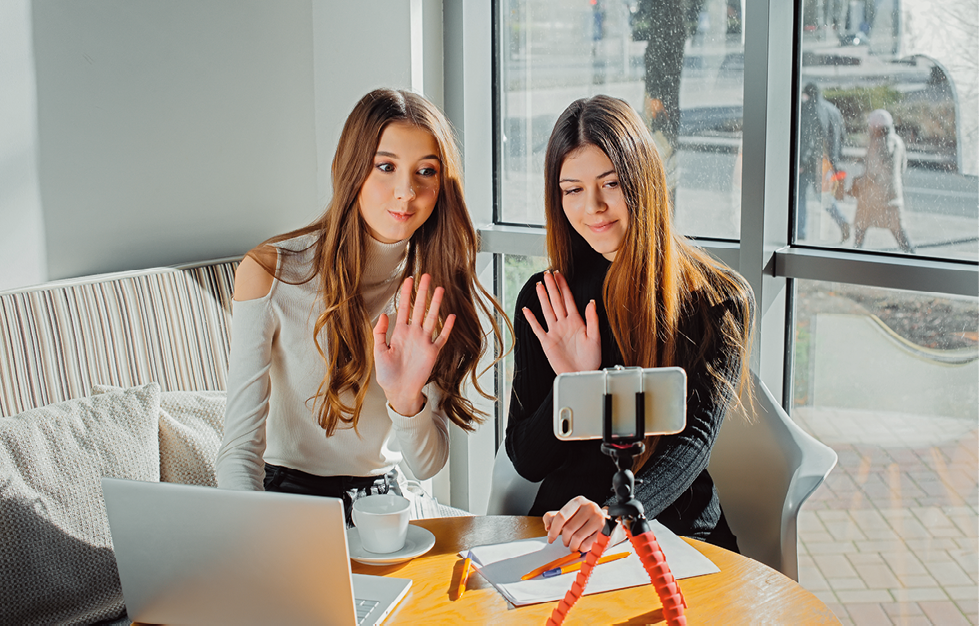 Fotografia. Duas meninas sentadas sobre cadeiras olhando para um celular, de frente para mesa em marrom-claro. Sobre ela, um celular bege na horizontal sobre um pedestal. A menina da esquerda, de cabelos longos ondulados em castanho e blusa de mangas compridas em bege-claro, com a mão esquerda acenando para o celular. A menina da direita, de cabelos longos em castanho, lisos e blusa de mangas compridas em preto. Ela está acenando com a mão direita, de frente para o celular. Ao fundo, janela vista parcialmente em branco e, mais ao fundo, rua vista parcialmente e pessoa caminhando para a esquerda.