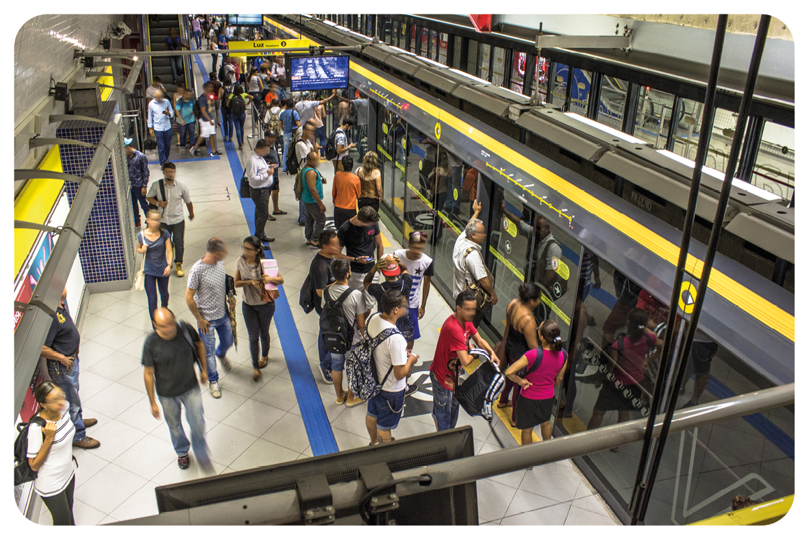Fotografia. Vista do alto de plataforma de metrô de cor bege-claro, linha em azul. À direita, proteção de vidro em preto, com detalhes acima em cinza e amarelo. De frente para a proteção, pessoas em pé. Na parte superior, pendurado, uma tela pequena preta e, à frente, tela em azul.