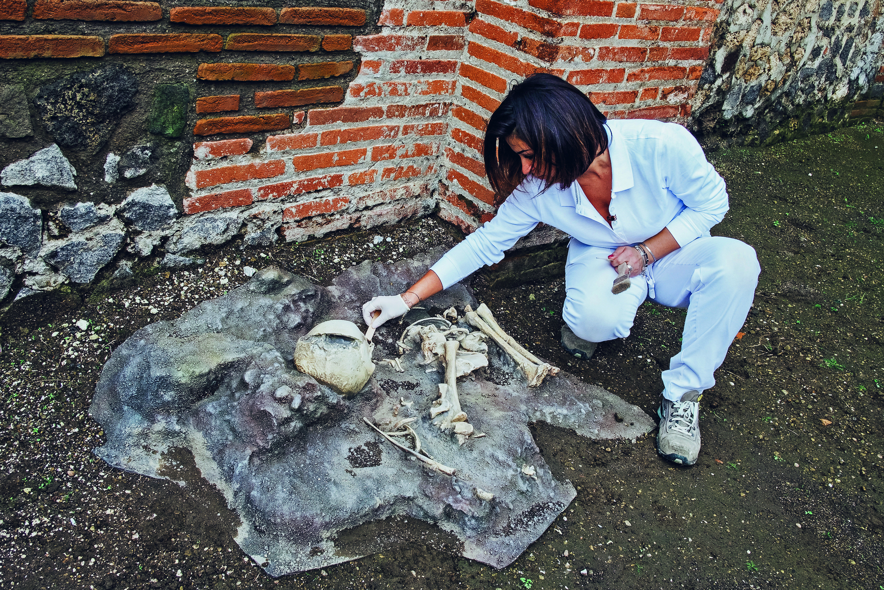 Fotografia. Mulher de roupas brancas usando luva cirúrgica analisa crânio e ossos humanos dispostos no chão de terra escura com paredes ao fundo.