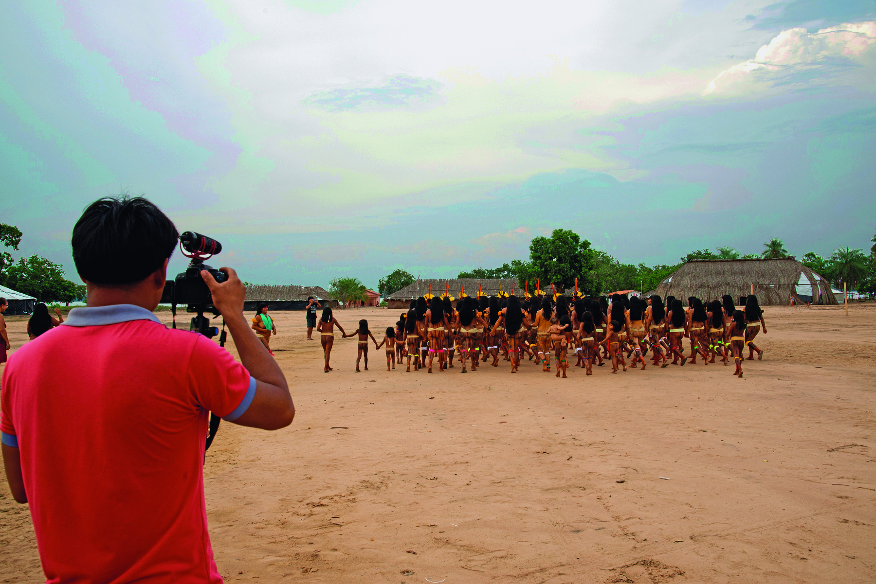 Fotografia. Em primeiro plano, homem indígena visto de costas, segurando uma câmera filmadora e, ao fundo, mulheres indígenas vistas de costas, de mãos dadas, no pátio de uma aldeia.