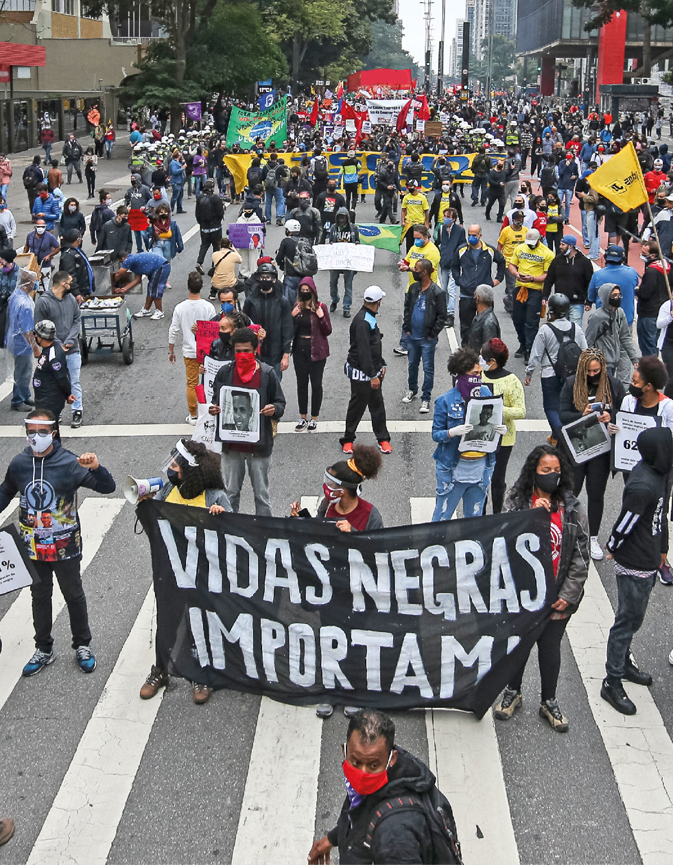 Fotografia. Multidão reunida em manifestação em uma grande avenida. Muitas pessoas utilizam máscara facial e seguram cartazes. Em uma faixa, em primeiro plano, lê-se: 'Vidas negras importam'.