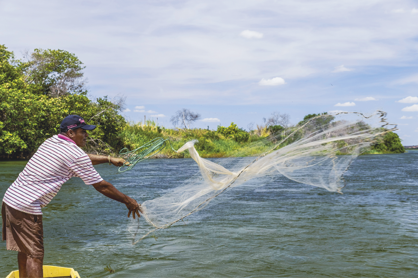 Fotografia. Homem de boné, bermuda e camisa listrada em cima de um pequeno barco em um rio jogando uma rede de pesca na água.