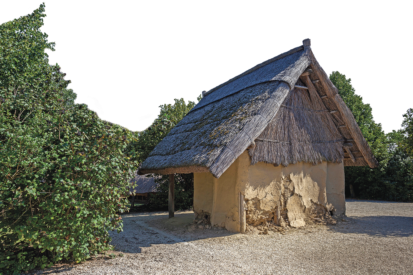 Fotografia. Pequena casa de barro com telhado triangular de palha em meio à vegetação.