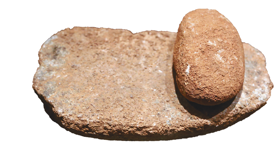 Fotografia. Dois artefatos de pedra: um comprido com a superfície achatada e lisa, e outro pequeno e arredondado. O objeto arredondado está posicionado à direita, sobre o objeto comprido.