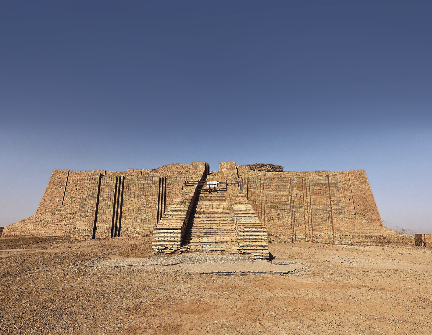 Fotografia. Construção quadrada de pedra vista de frente com uma grande escadaria na parte frontal, em um campo aberto coberto de areia. Ao fundo, céu azul.