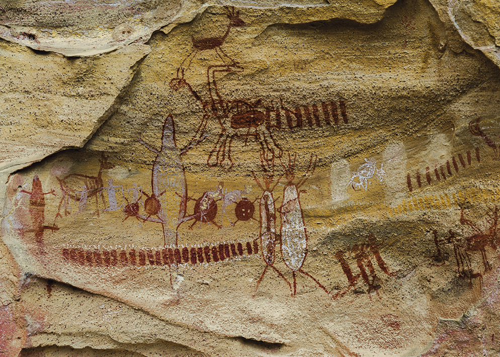 Fotografia. Pintura rupestre nas cores vermelha, branca e amarela, com formas de animais e de seres humanos, em parede de uma caverna.