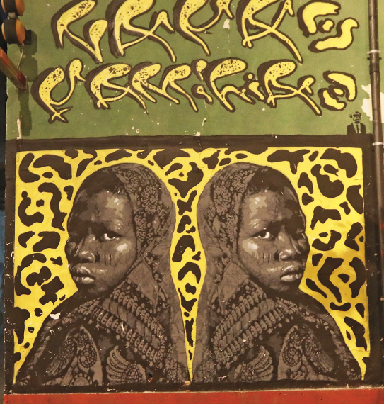 Fotografia. Grafite representando uma menina negra com véu na cabeça desenhada duas vezes, uma de costas para outra. Ao fundo, desenhos assimétricos em preto e amarelo. Em cima, letras estilizadas em amarelo sobre um fundo verde.