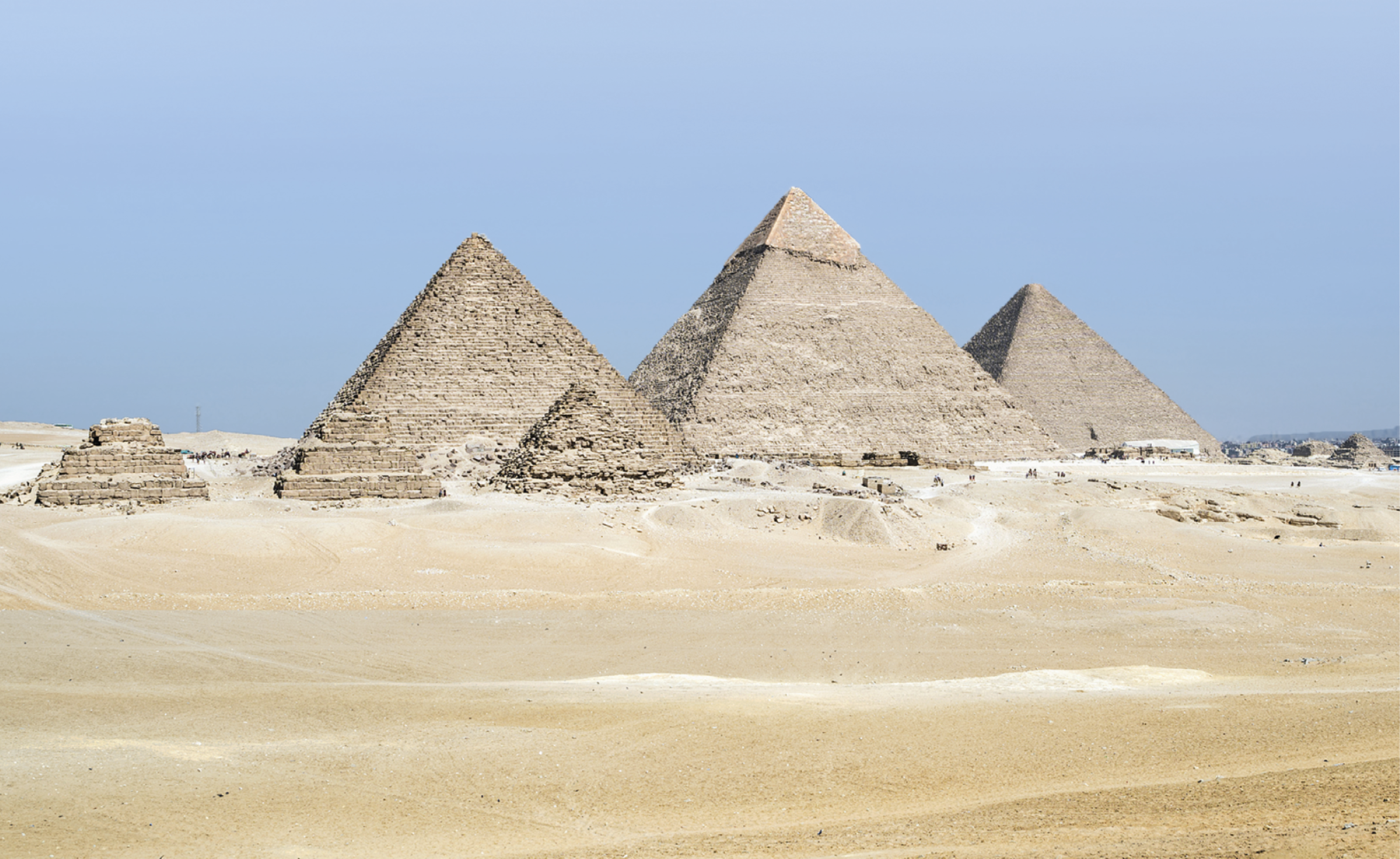 Fotografia. Vista de pirâmides egípcias em um campo aberto. Em primeiro plano, terreno arenoso e três pirâmides menores, à esquerda. Em segundo plano, três pirâmides maiores, ao centro. Ao fundo, céu azul.