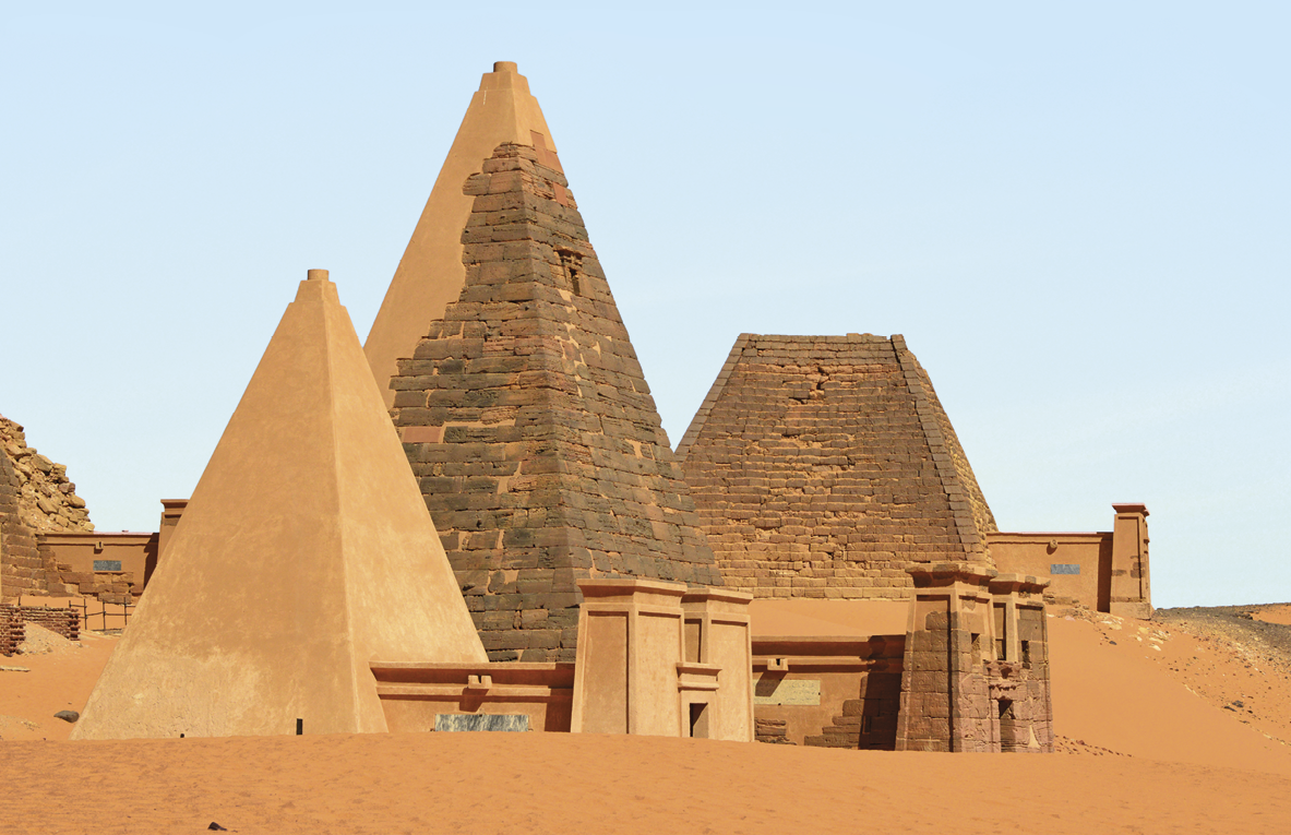 Fotografia. Destaque para pirâmides altas de base mais estreita e pontiagudas e duas construções menores à frente, com colunas de base quadrada, que servem como portal de entrada das pirâmides. O solo é arenoso. Ao fundo, céu azul.