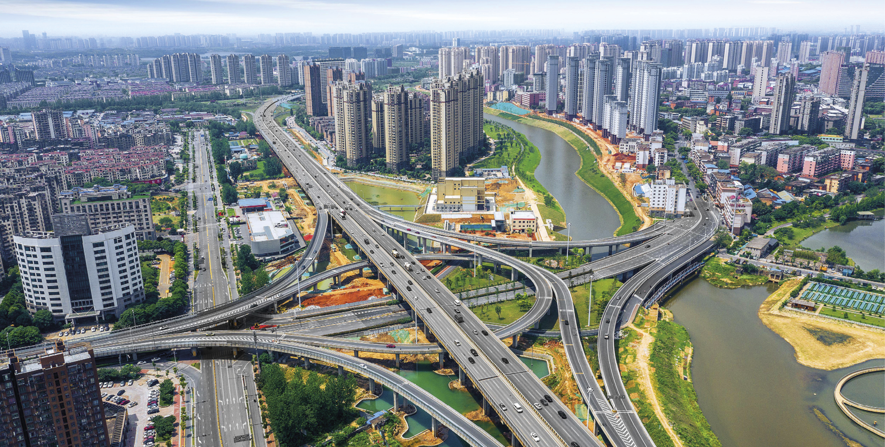 Fotografia. Vista aérea de uma cidade moderna, com destaque para várias avenidas, pontes e viadutos interconectados. À direita, um rio cujas margens direita e esquerda são ligadas por diversas pontes. Ao fundo, grandes prédios.