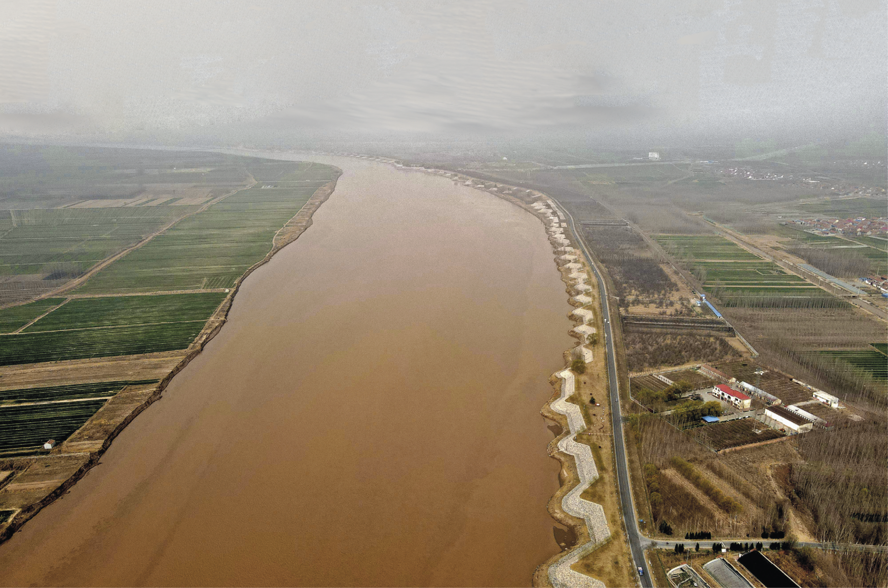 Fotografia. Vista aérea de um rio de cor barrenta com plantações nas duas margens.