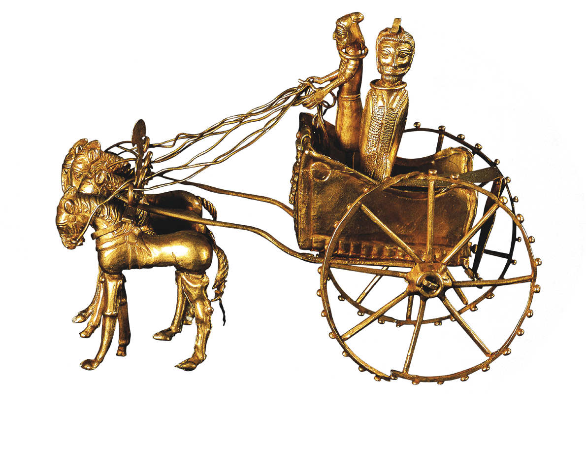 Fotografia. Escultura de ouro de uma carruagem com dois homens dentro, um que segura as rédeas e veste apenas um pano amarrado à cintura, e outro vestido com um manto com vários adornos, sendo puxada por quatro cavalos.