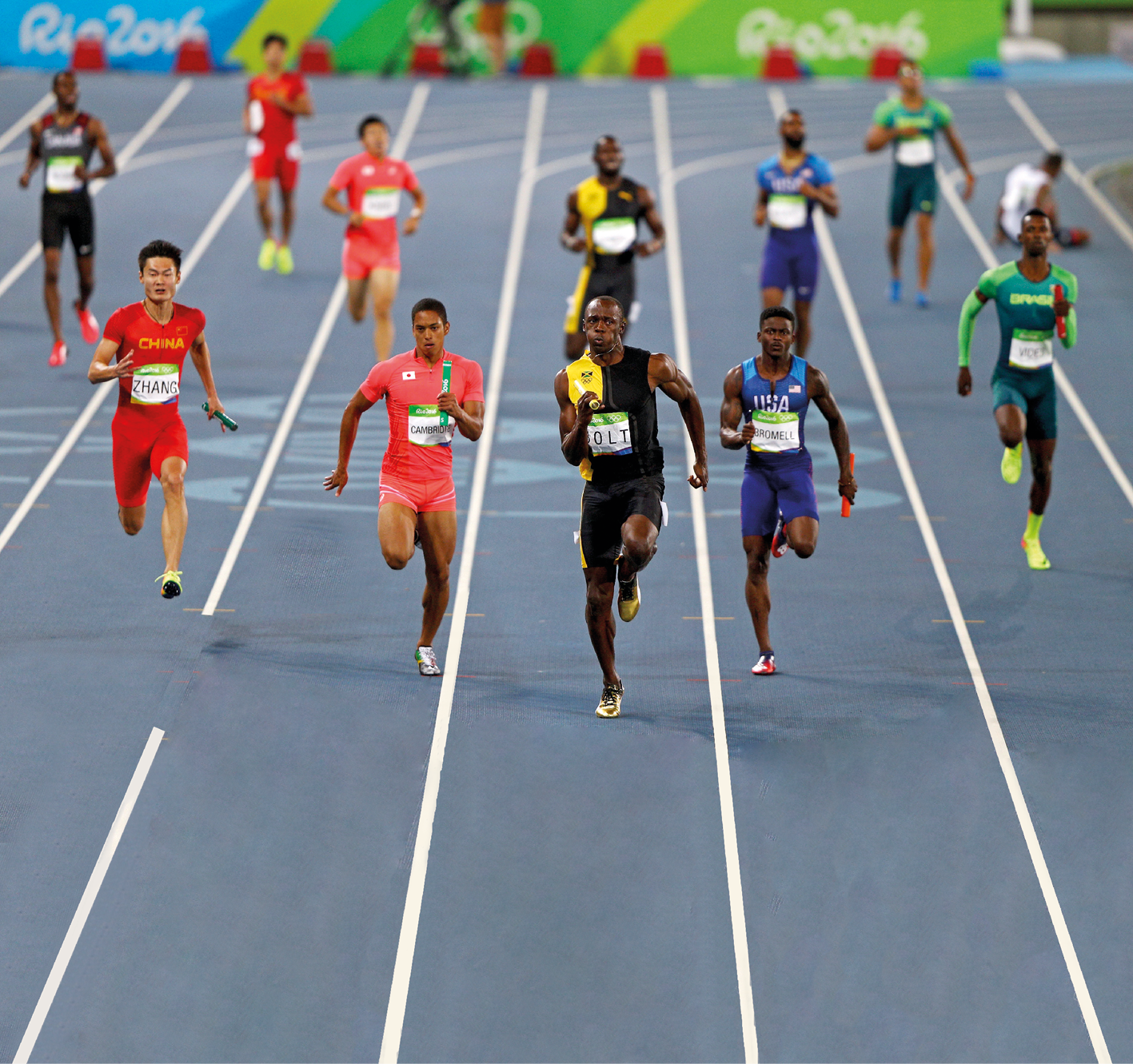 Fotografia. Em primeiro plano, homens correndo com bastões nas mãos em raias de uma pista de atletismo. Em segundo plano, nas mesmas raias e com os mesmos uniformes dos homens da frente, homens diminuindo a velocidade. Ao fundo, placas coloridas com a inscrição "Rio 2016".