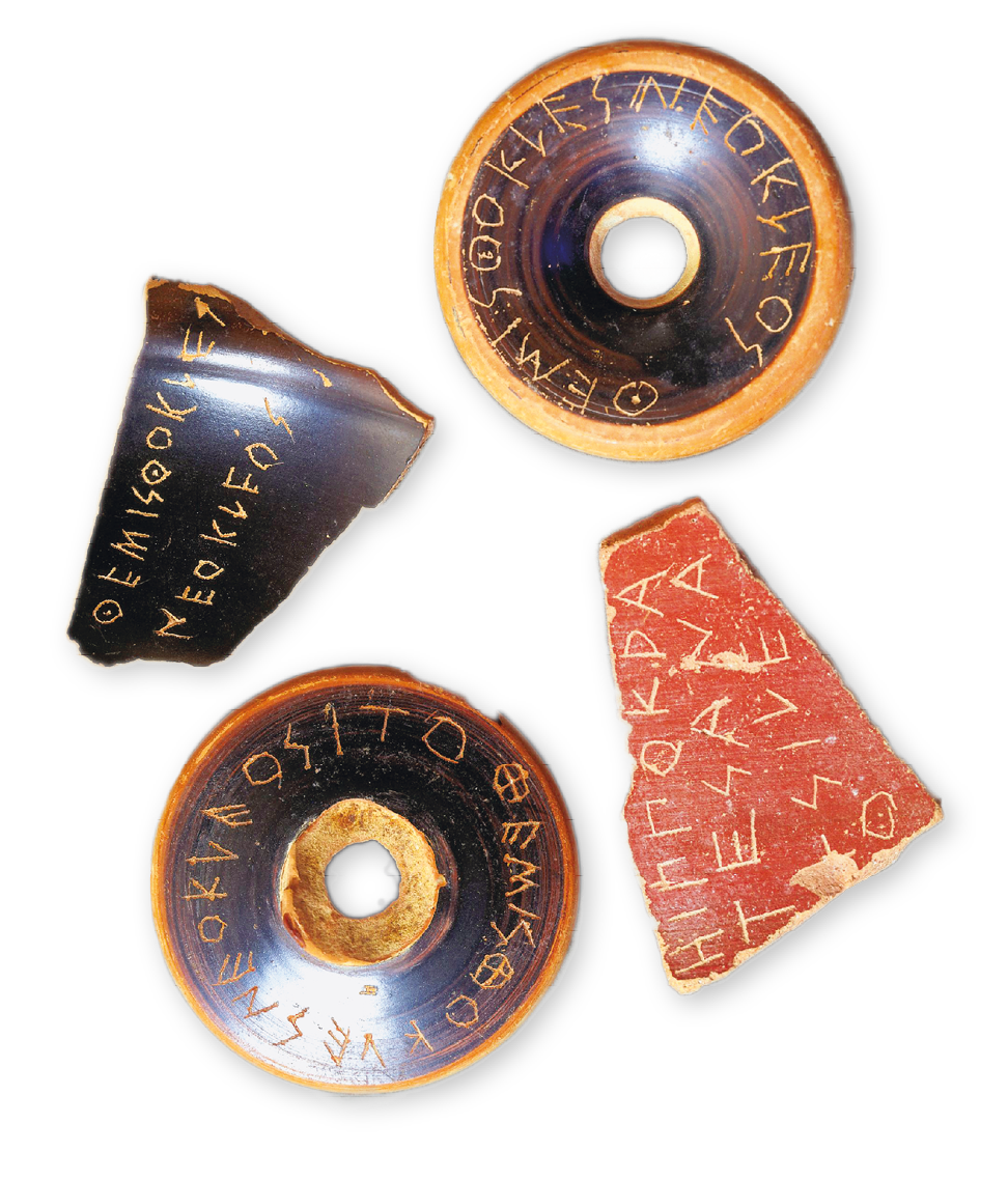 Fotografia. Quatro artefatos de cerâmica, dois arredondados com um furo no meio e dois de formato irregular, semelhante a um caco. Todos possuem inscrições em grego.