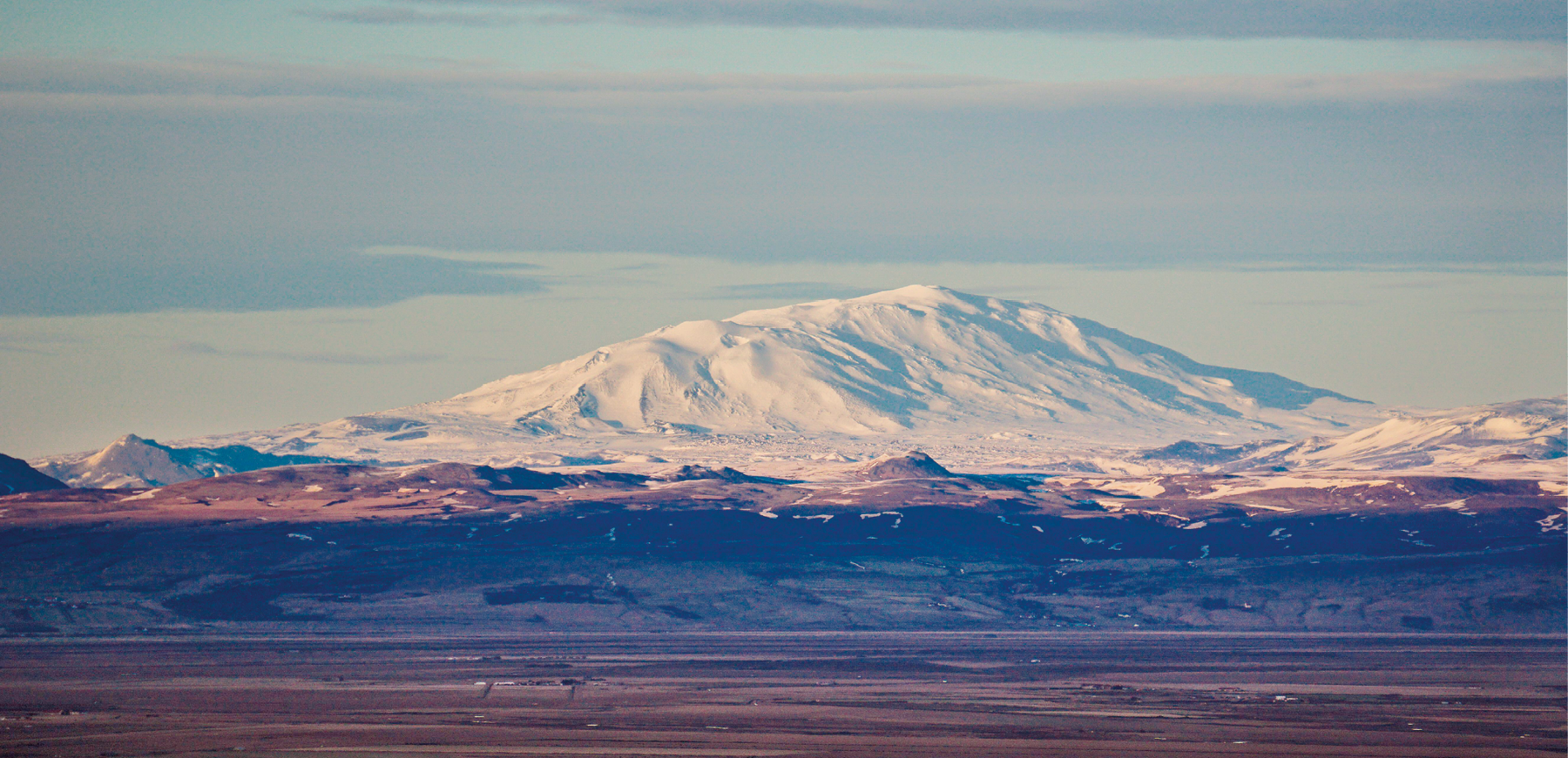 Fotografia. Um grande vulcão coberto de neve, ao fundo. Em segundo plano, montes menores sem neve e, em primeiro plano, campo aberto.