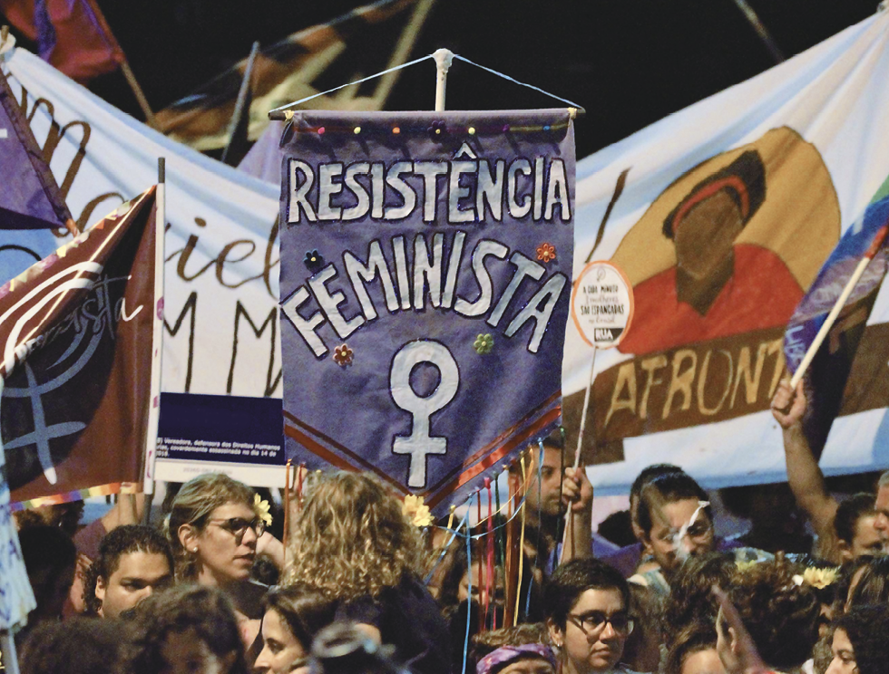 Fotografia. Mulheres em uma manifestação, com muitos cartazes. Em destaque, no centro uma flâmula lilás, onde se lê com letras brancas: "Resistência feminista" e, abaixo, o símbolo de um círculo com uma cruz embaixo.