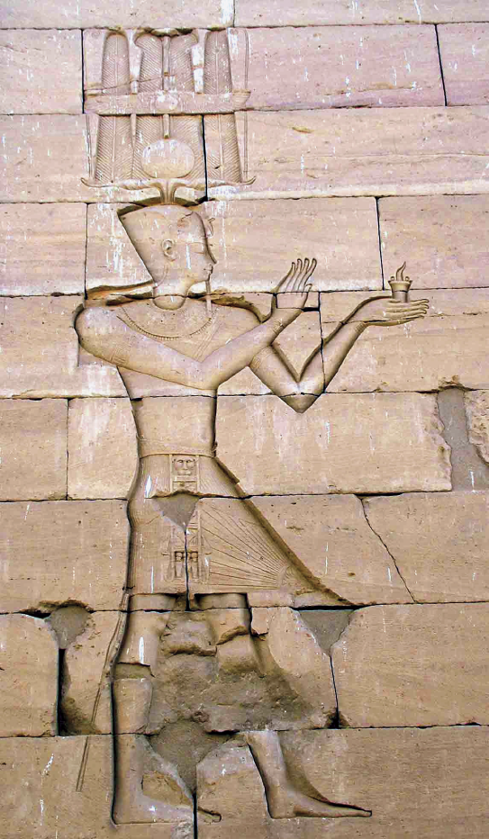 Fotografia. Parede feita de blocos de pedra com a figura de um homem entalhada em baixo-relevo. O homem, de perfil, usa um chapéu de base quadrada, saia e colar egípcios ergue as mãos, segurando um objeto em uma delas.