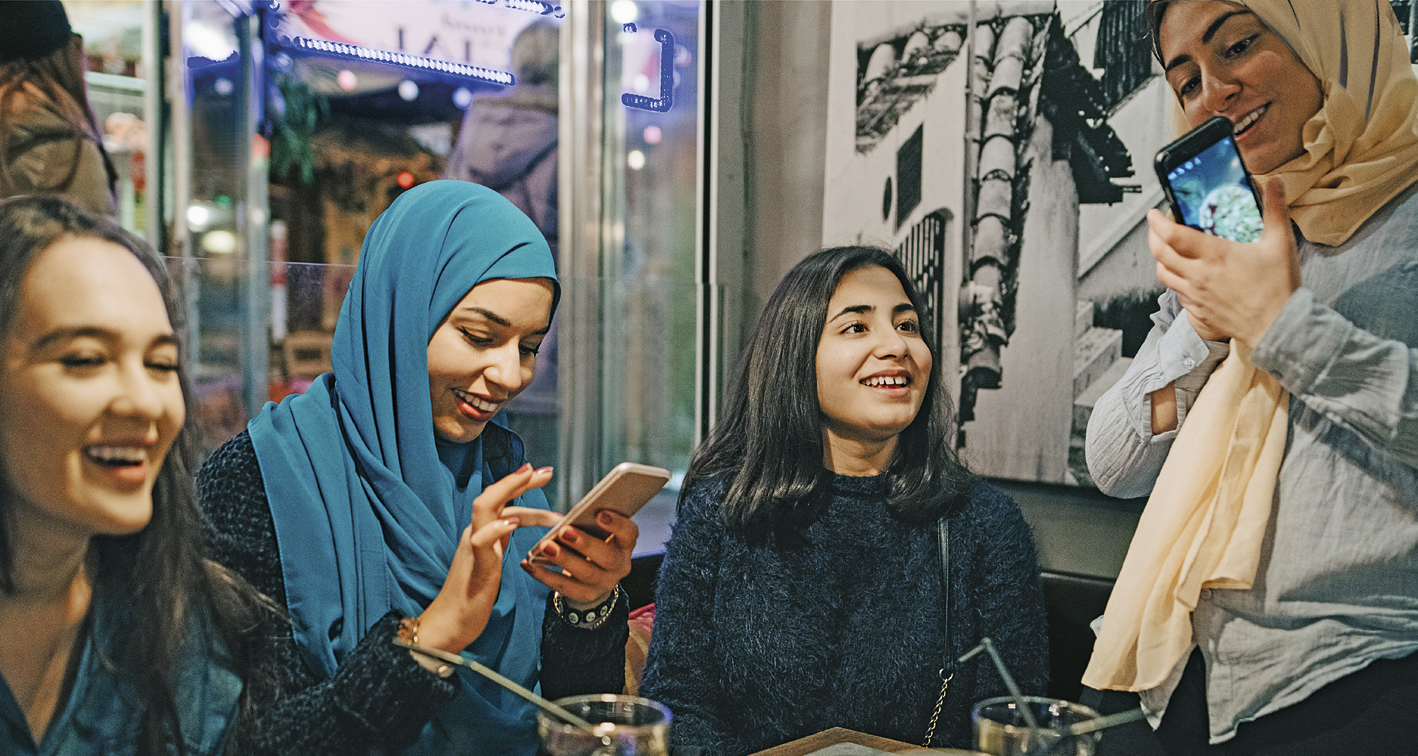 Fotografia. Quatro mulheres reunidas. Duas delas usam hijabes e seguram celulares. Elas estão sorrindo.
