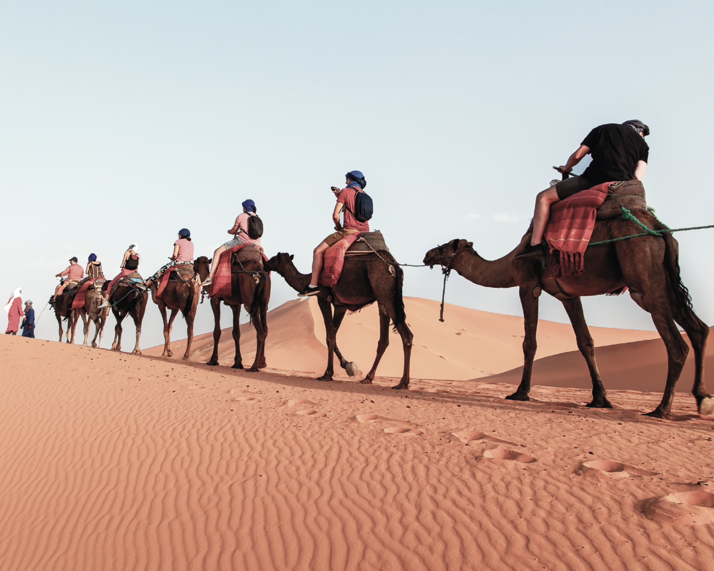 Fotografia. Fila de pessoas com camisetas, bermudas e mochilas, usando lenços na cabeça, montadas em camelos, que andam pelo deserto.