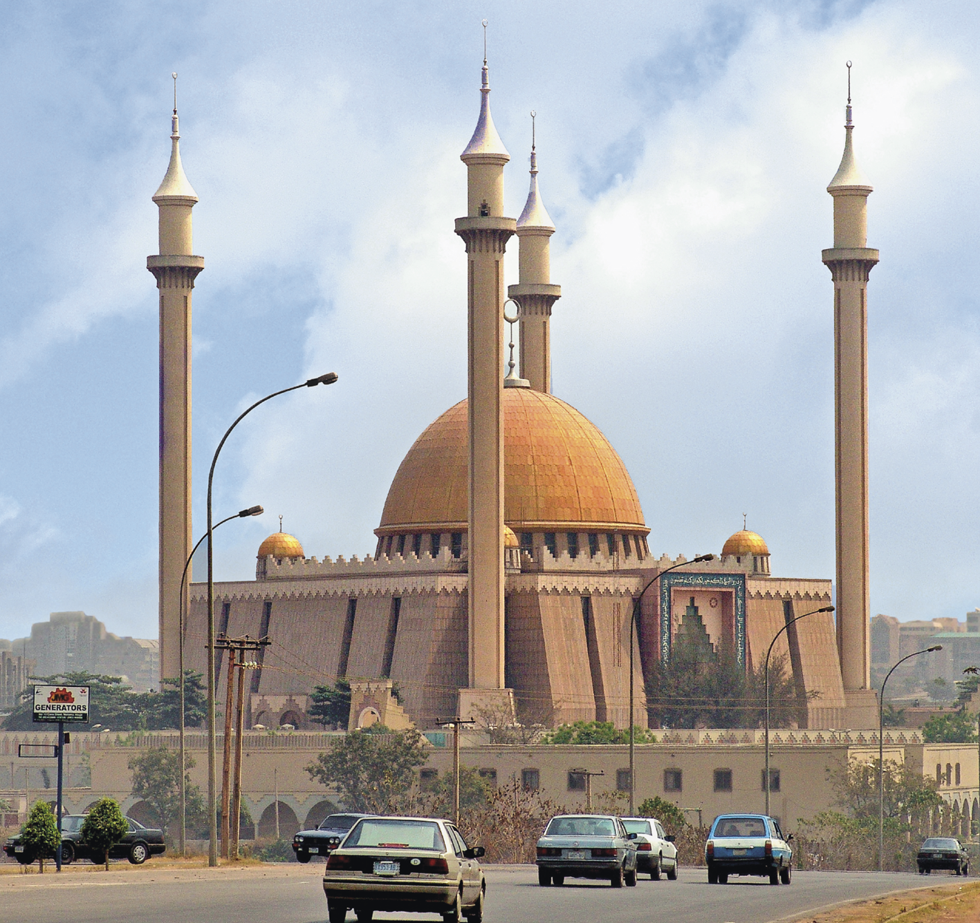 Fotografia. Ao fundo, uma mesquita tradicional, rodeada por uma muralha e construções pontudas. À frente, carros em uma rodovia.