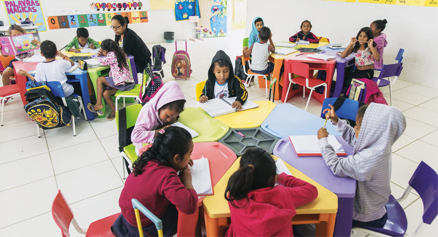 Fotografia. Sala de aula com três mesas coloridas, de forma hexagonal, e crianças sentadas em volta delas. As crianças escrevem em cadernos e, próximo a uma das mesas, à esquerda, uma professora sentada ajuda um grupo de alunos.
