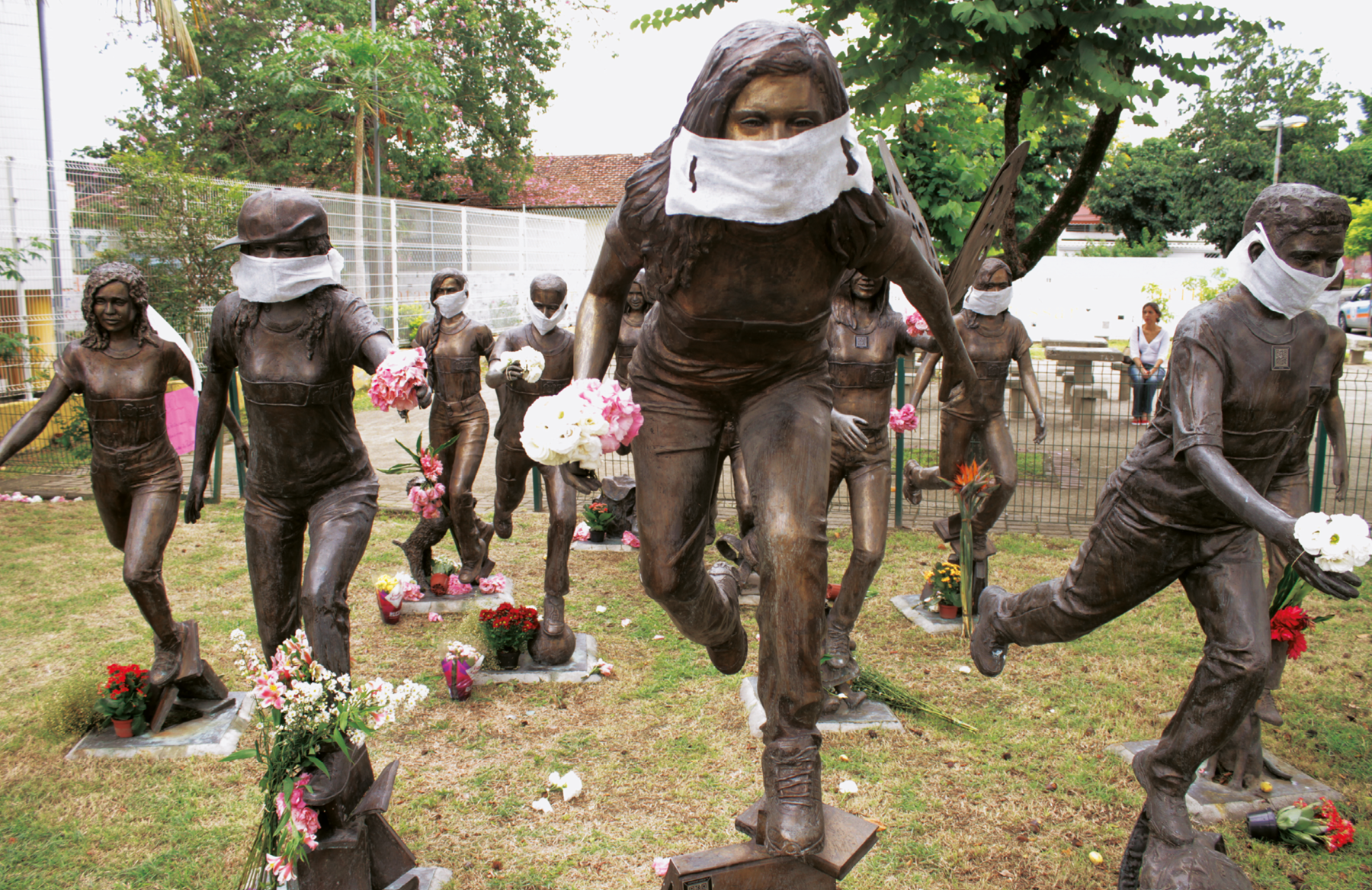 Fotografia. Esculturas de jovens em posição de corrida. Em algumas delas foram colocadas máscaras de proteção facial. Elas seguram flores coloridas nas mãos. Aos seus pés, há vasos e buquês de flores.