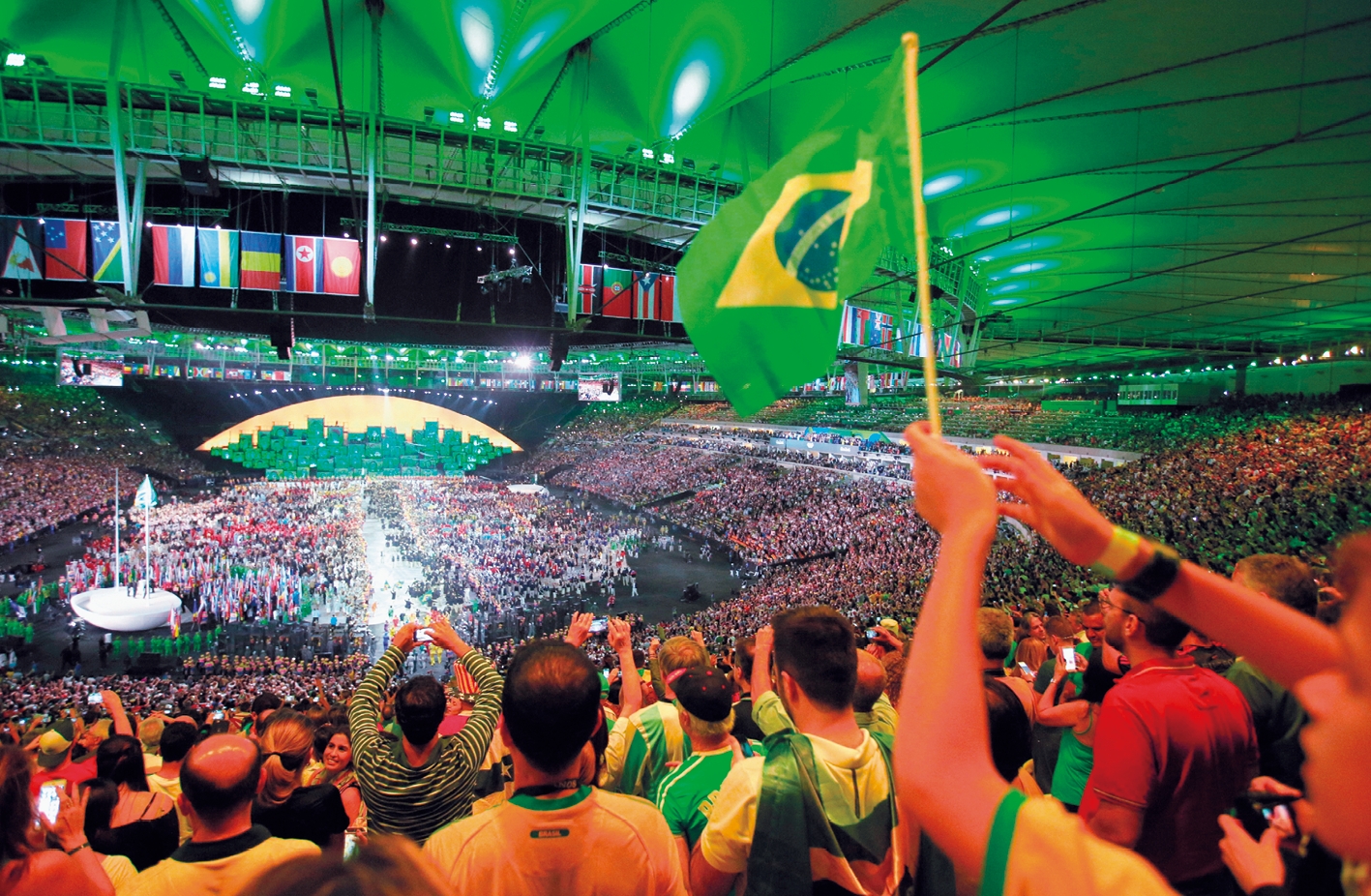 Fotografia. Vista do interior de um estádio de futebol coberto, com diversas pessoas com camisas do Brasil ocupando as arquibancadas e, ao centro, um espetáculo acontecendo. No alto, bandeiras de vários países enfileiradas.