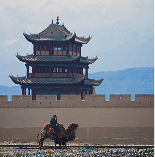 Fotografia. Ao fundo, uma torre oriental no estilo pagode. Ela é protegida por uma muralha, que aparece em segundo plano. Em primeiro plano, uma pessoa montada em um camelo.