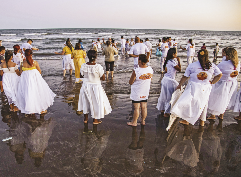 Fotografia. Diversas pessoas aparecem de costas e vestidas de branco em uma faixa de areia. Algumas, em primeiro plano, estão à beira do mar, enquanto outras, mais ao fundo, com água à altura das canelas.