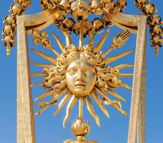Fotografia. Detalhe dourado de um portão com a cabeça de um homem de cabelos encaracolados, da qual saem raios de sol.