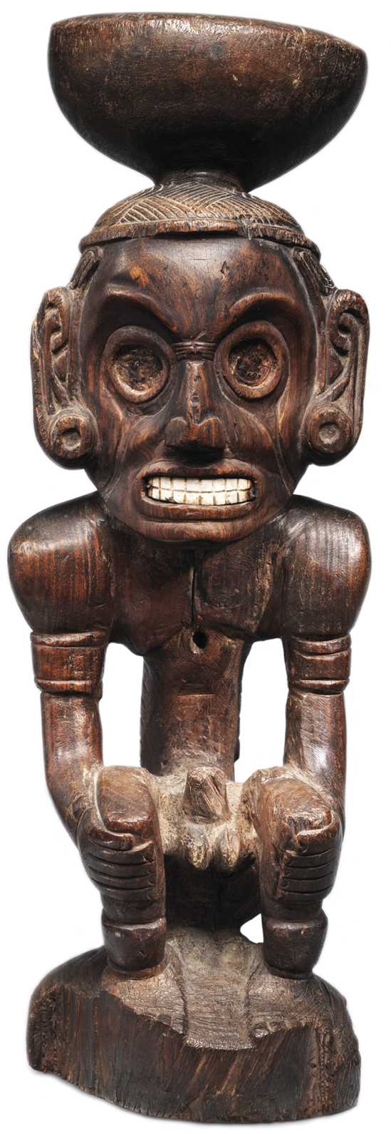 Fotografia. Escultura de madeira de um homem nu com os dentes cerrados sentado com as mãos nos joelhos e um pote na cabeça.