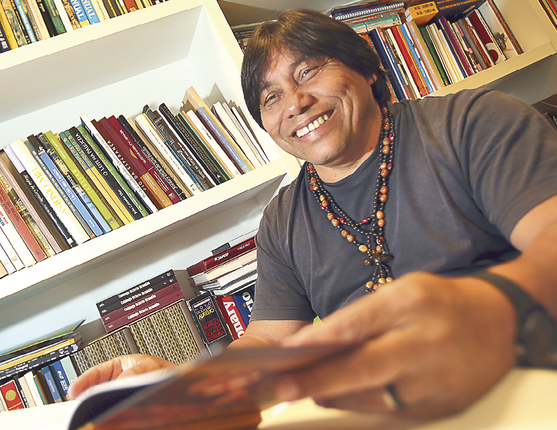 Fotografia. Homem indígena de cabelos pretos curtos, com colares de miçangas pretas e laranjas. Ele está sorrindo sentado diante de uma mesa e segura um livro aberto. Ao fundo, uma estante cheia de livros.